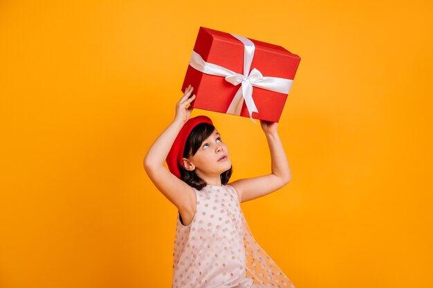 Criança morena interessada adivinhando o que há no presente. menina com chapéu vermelho, segurando o presente na parede amarela.