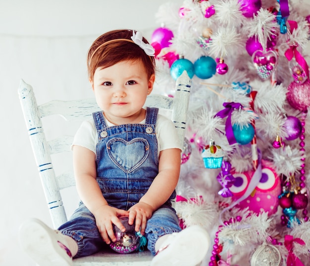 Criança pequena bonita senta na cadeira antes da árvore de Natal