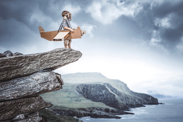 Criança vestida de aviador com avião de papelão em um penhasco sonha com uma aventura.
