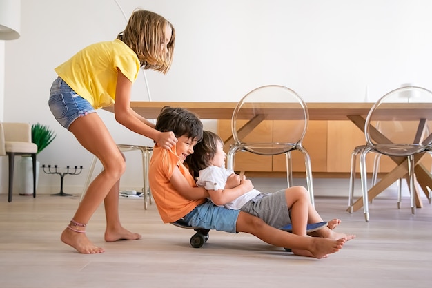 Crianças alegres brincando com o skate em casa. Adorável menina loira empurrando seus dois irmãos brincalhões. Crianças felizes andando a bordo e se divertindo. Infância, atividade de jogo e conceito de fim de semana