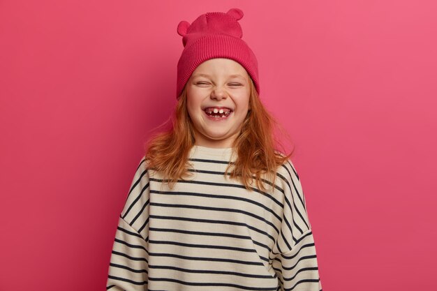 Crianças e o conceito de felicidade. A ruiva alegre ri de algo engraçado, usa chapéu rosa com orelhas e um suéter listrado solto, sorri brilhantemente, tem dentes faltando, modelos dentro de casa.