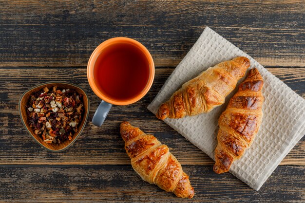 Croissant com chá na xícara, ervas secas planas leigos na toalha de madeira e cozinha