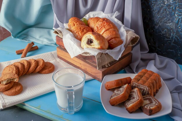 Croissants com creme de chocolate, torta de baunilha e biscoitos com um copo de leite na mesa de madeira azul.