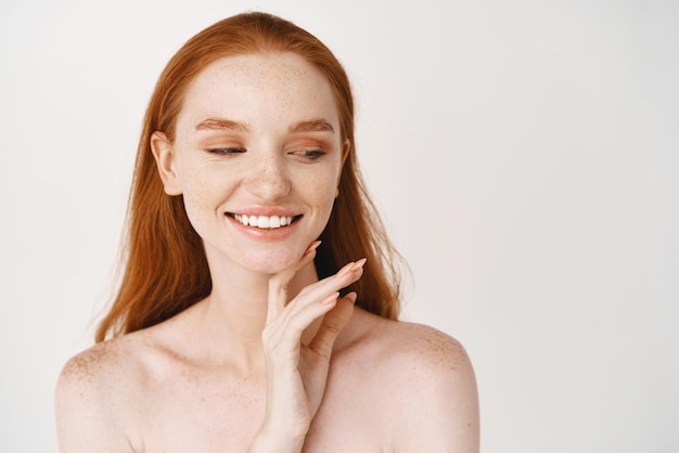 Cuidados com a pele closeup de mulher ruiva com pele macia pálida sorrindo dentes brancos e tocando rosto limpo sem maquiagem olhando de lado em pé sobre fundo branco nu