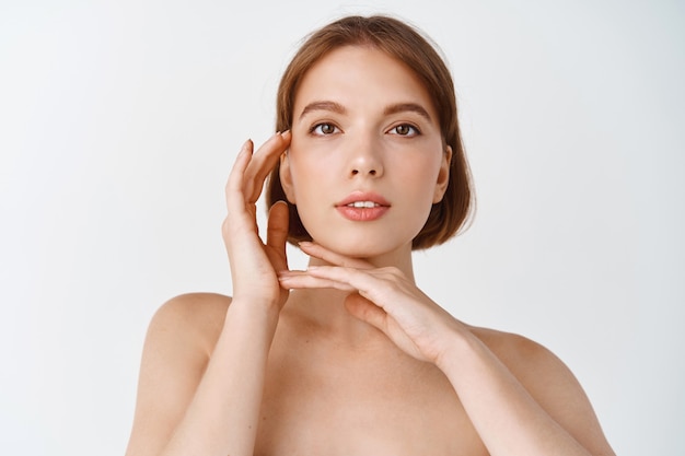 Cuidados com a pele e beleza. Jovem natural em pé nua, tocando o rosto brilhante. Menina mostrando efeito de produto para a pele, pele limpa e fresca sem manchas, parede branca