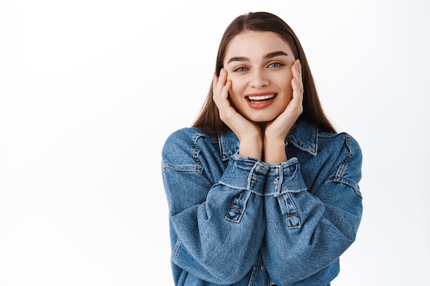 Foto grátis cuidados com a pele e beleza. linda e feliz adolescente tocando a pele natural pura, limpeza facial fresca, sorrindo satisfeita, encostada na parede branca