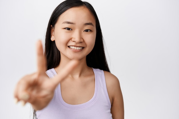 Cuidados com a pele e bem-estar feminino. Menina asiática feliz sem maquiagem, mostrando o símbolo V da paz, sorrindo e parecendo alegre em branco sobre branco