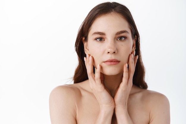 Cuidados com a pele. mulher jovem e bonita com maquiagem natural, tocando a pele do rosto hidratada e nutrida, de mãos dadas nas bochechas, nua contra um fundo branco