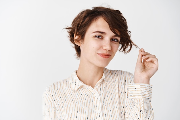 Cuidados com o cabelo e cuidados com a pele linda mulher de blusa brincando com fio de cabelo e olhando para a câmera com sorriso em pé sobre fundo branco