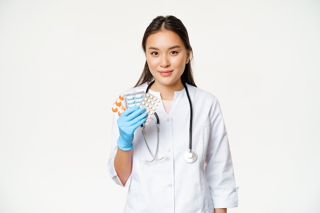 Cuidados de saúde e conceito médico. Médica asiática mostrando pílulas, vitaminas em luvas estéreis de borracha, de uniforme sobre fundo branco