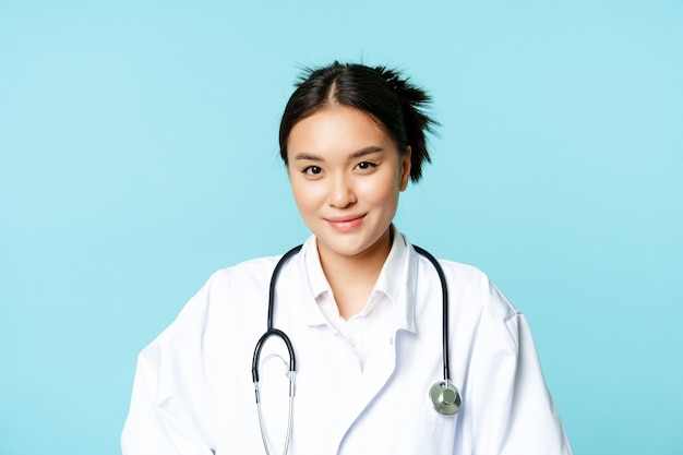 Cuidados de saúde e conceito médico. Médica coreana, enfermeira de uniforme, sorrindo e parecendo prestativa, fundo azul