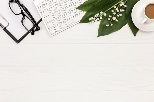 Óculos, prancheta, teclado, flor e folhas com xícara de café na mesa de escritório