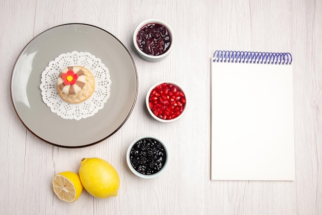 Foto grátis cupcake de vista superior e prato de geleia de cupcake apetitoso no guardanapo de renda ao lado das tigelas de caderno branco com sementes de geleia de romã e limão na mesa