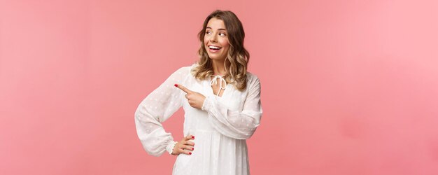 Curiosa e otimista linda mulher loira aproveitando o verão se sentindo alegre apontando olhando para a esquerda com expressão entusiasmada interessada usar vestido branco fofo em pé fundo rosa