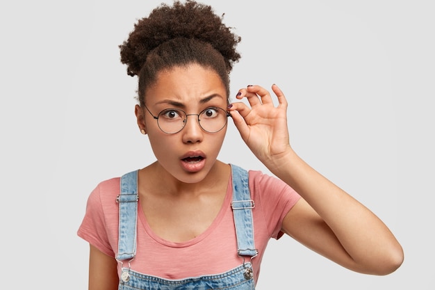 Curiosa e surpresa jovem de pele escura olha escrupulosamente através de óculos redondos, tem cabelo preto cacheado penteado em coque, olha atento para alguma coisa, usa macacão jeans com camiseta