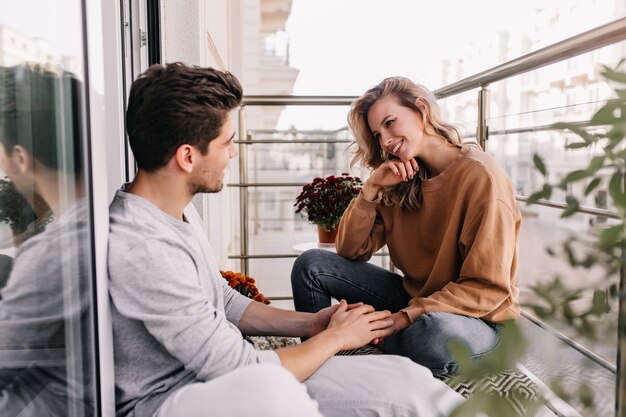 Curiosa garota caucasiana falando com um amigo no terraço. Sonhadora jovem sentada na varanda com o namorado.