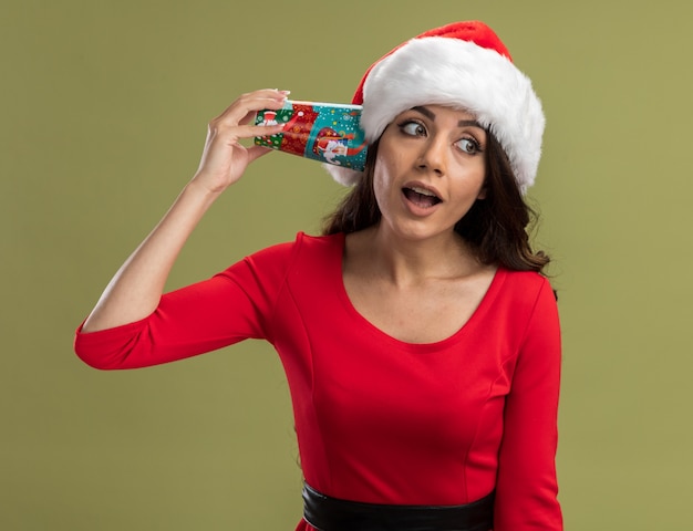 Curiosa jovem bonita com chapéu de papai noel segurando a xícara de café de natal perto da orelha, ouvindo uma conversa olhando para o lado