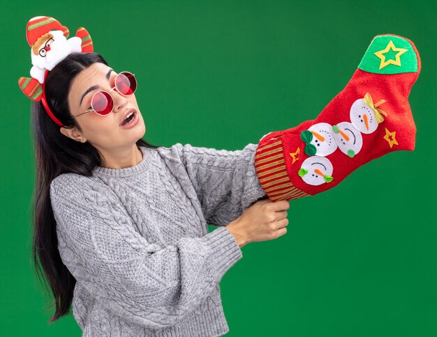 Curiosa jovem caucasiana usando bandana de papai noel com óculos segurando uma meia de natal olhando para ela colocando a mão dentro dela isolada na parede verde