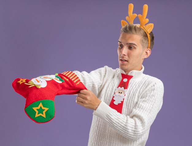 Curioso, jovem, bonito, usando uma bandana de chifres de rena e gravata de papai noel segurando e olhando a meia de natal, colocando a mão dentro dela, isolada na parede roxa