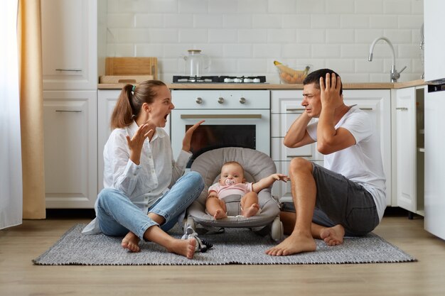 Curto interior discutindo casal sentado no chão da cozinha, esposa gritando alto, marido cobrindo as orelhas com as palmas das mãos, família posando com bebê infantil na cadeira de balanço.