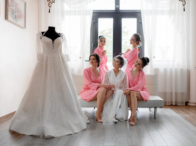 Damas de honra sorriu felizes com a noiva está olhando para o vestido de casamento na sala de luz, preparação do casamento