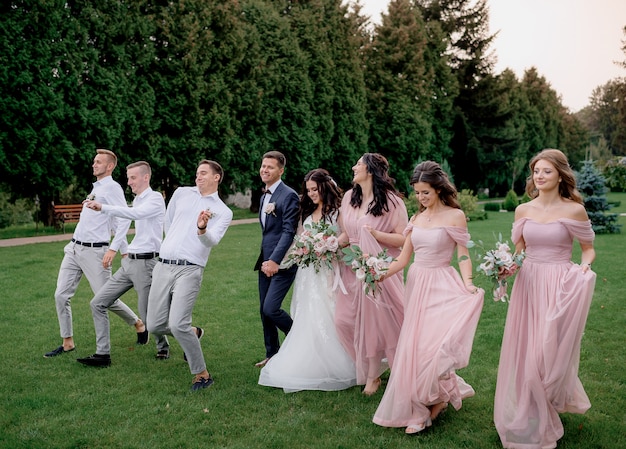 Damas de honra vestidas com vestidos cor de rosa, os melhores homens e o casal de noivos estão felizes andando no quintal verde