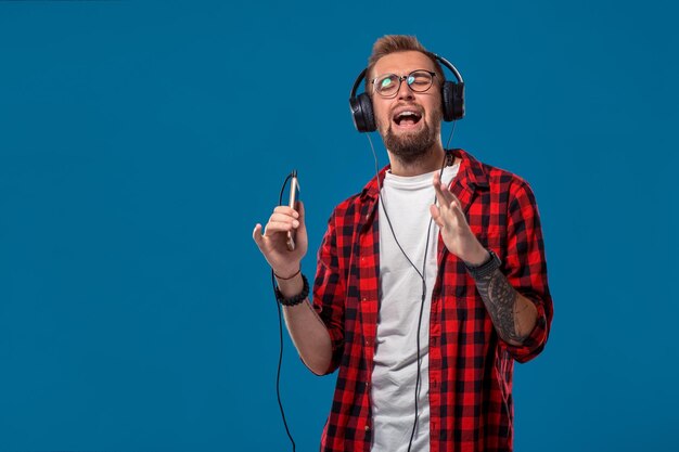 Dança bonito jovem elegante em fones de ouvido segurando o MP3 Player e dançando em pé contra um fundo azul. Conceito emocional e de pessoas: jovem barbudo de camisa quadriculada. Estilo hipster.