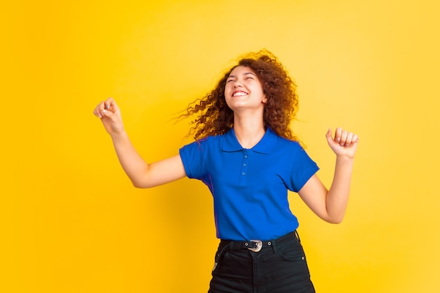 Dançando com cabelos voando. Retrato de menina adolescente caucasiano em fundo amarelo do estúdio. Lindo modelo feminino encaracolado. Conceito de emoções humanas, expressão facial, vendas, anúncio, educação. Copyspace.