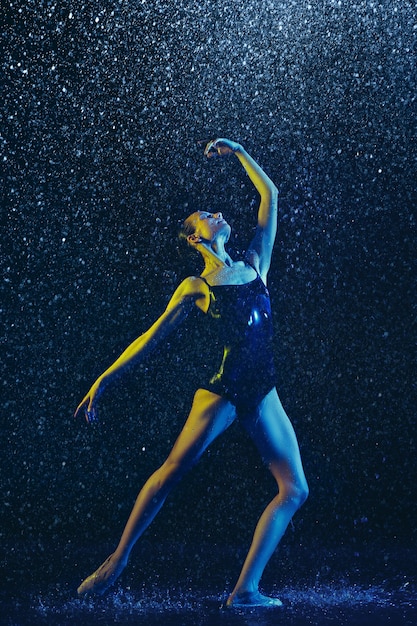 Dançarina de balé feminino jovem realizando sob spray e gotas de água. Modelo caucasiano dançando em luzes de néon. Mulher atraente. Conceito de balé e coreografia contemporânea. Foto de arte criativa.