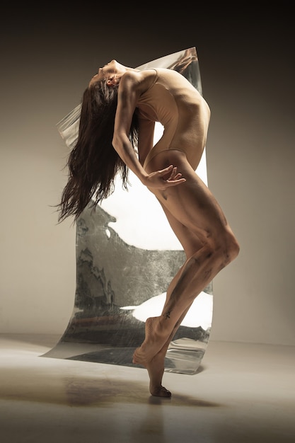 Dançarina de balé moderno jovem e elegante na parede marrom com o espelho e reflexos de ilusão na superfície