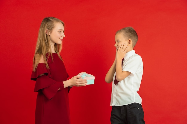 Dando coração. celebração do dia dos namorados, crianças brancas felizes e fofas isoladas no fundo vermelho do estúdio. conceito de emoções humanas, expressão facial, amor, relações, férias românticas.