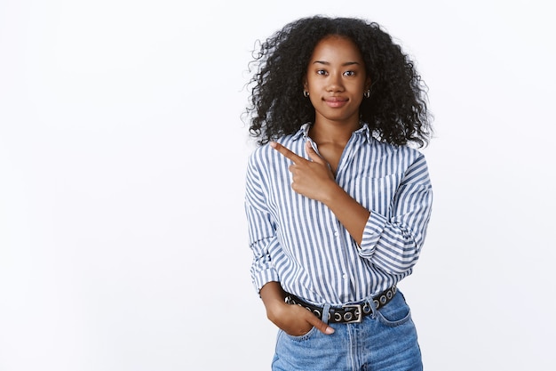 De aparência amigável, determinada satisfeita, alegre, jovem afro-americana cliente posando com uma parede branca apontando para o lado, indicando o dedo indicador esquerdo sorrindo, escolha escolher variante