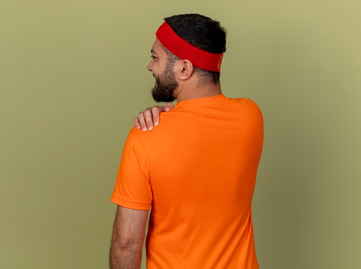 Foto grátis de pé, de costas para a câmera, jovem desportivo usando fita para a cabeça e pulseira, segurando o ombro dolorido isolado em fundo verde oliva