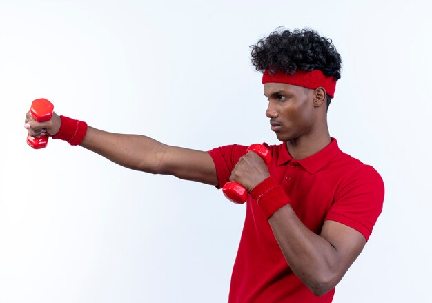 De pé em vista de perfil, jovem afro-americano esportivo usando bandana e pulseira segurando halteres isolados no branco