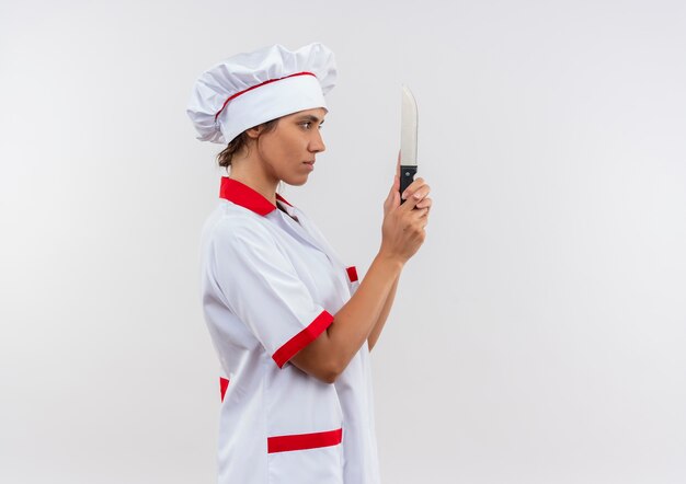 De pé na vista de perfil jovem cozinheira vestindo uniforme de chef segurando e olhando a faca com espaço de cópia
