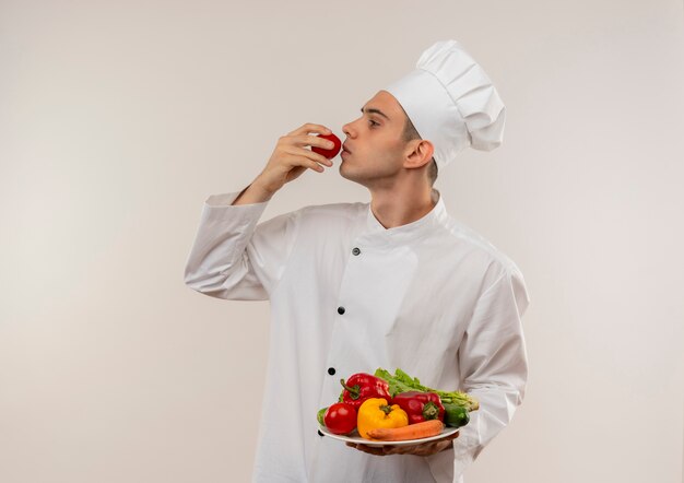 De pé na vista de perfil, jovem cozinheiro masculino vestindo uniforme de chef, segurando vegetais no prato e cheirando tomate na mão com espaço de cópia