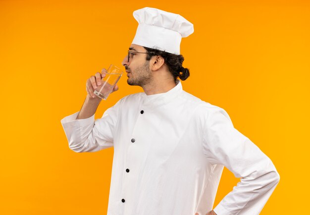 De pé na vista de perfil, jovem cozinheiro vestindo uniforme de chef e óculos, bebeu água isolada na parede amarela