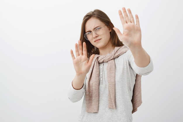 Dê um passo para trás, você me incomoda. Mulher intensa e fofa descontente de óculos com cabelo castanho curto curvado para trás, levantando as palmas das mãos em defesa, tentando proteger os olhos da luz do sol ou da lanterna sobre a parede cinza