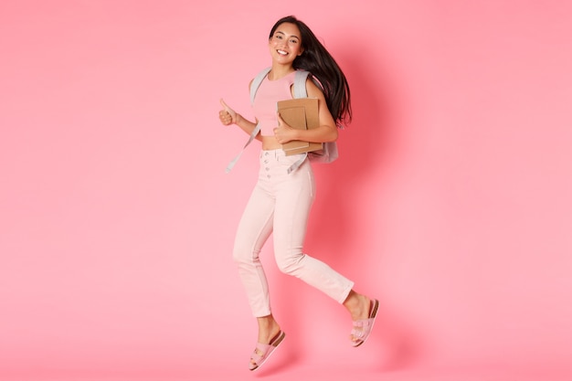 De volta ao conceito de escola, educação e estilo de vida. comprimento total de uma aluna asiática sorridente feliz, garota da faculdade pulando com cadernos e mochila, mostrando o polegar para cima em aprovação, parede rosa