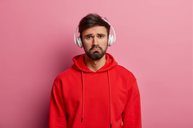 Decepcionado frustrado homem infeliz tenta se divertir com música, tem expressão facial melancólica, usa fones de ouvido nas orelhas, veste um moletom com capuz vermelho, isolado sobre parede rosa pastel.