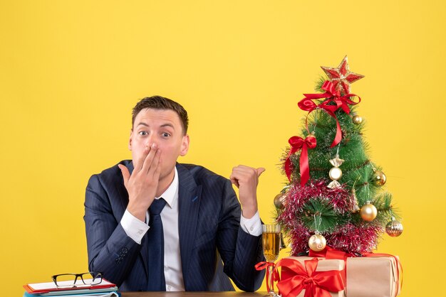 Dedo de homem espantado de vista frontal apontando a árvore de natal sentado à mesa perto da árvore de natal e presentes em fundo amarelo