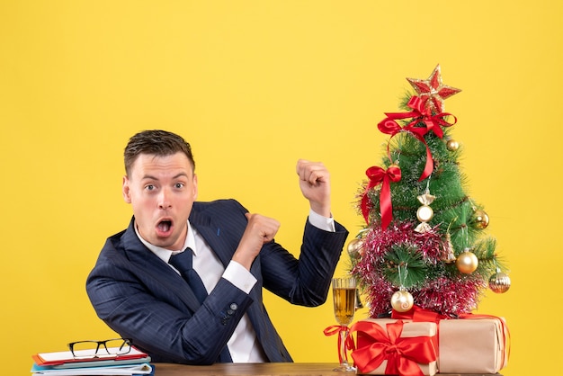 Dedo de homem espantado de vista frontal apontando para trás sentado à mesa perto da árvore de natal e presentes em fundo amarelo