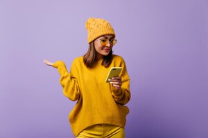 Foto grátis deliciosa agradável aluna da europa olhando com surpresa para sms em seu telefone. menina com chapéu quente amarelo e óculos coloridos posando para retrato isolado