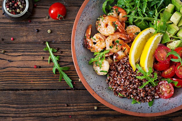 Deliciosa tigela de Buda saudável com camarões, tomate, abacate, quinoa, limão e rúcula na mesa de madeira. Comida saudável. Vista do topo. Postura plana.