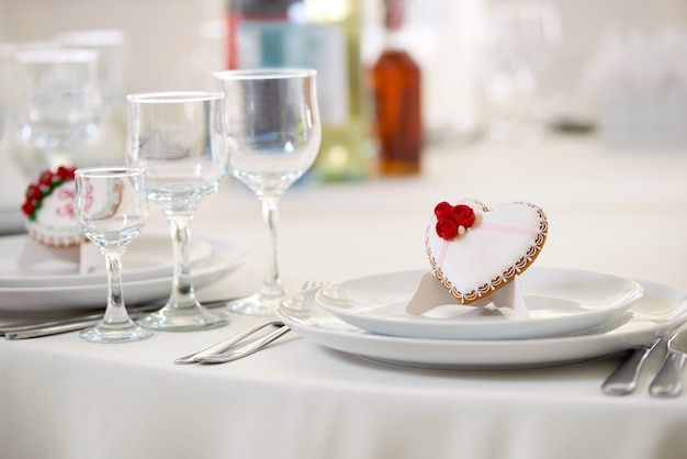 Delicioso biscoito coberto com esmalte branco doce e decorado com pequenas rosas vermelhas e pequenas pérolas brancas fica na mesa, servido com taças de vinho. Boa decoração para mesa festiva de casamento.