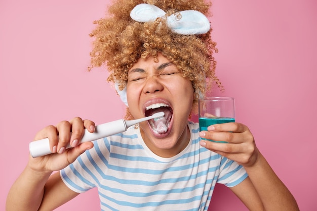 Foto grátis dentes saudáveis e conceito de cuidado mulher de cabelos cacheados alegre escova os dentes com escova de dentes elétrica segura copo de enxaguante bucal azul cuida da cavidade oral usa faixa de cabeça e camiseta listrada