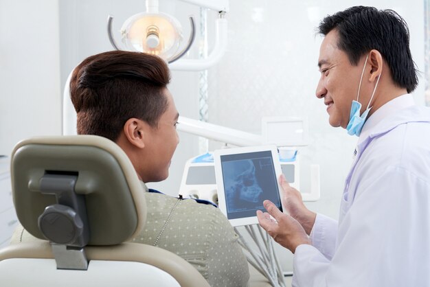 Dentista masculina asiática, demonstrando a imagem de raio-x da mandíbula no tablet para o paciente