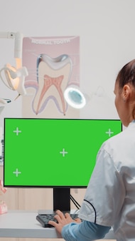 Dentista usando computador com tela verde horizontal em exibição. especialista olhando para o monitor com chroma key e gabarito isolado para cuidados bucais enquanto assistia ao uso de equipamentos odontológicos