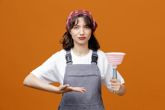 Desagradado jovem limpador feminino vestindo uniforme e bandana mostrando êmbolo apontando para ele com a mão olhando para câmera isolada em fundo laranja