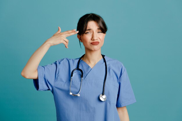 Desagradado mostrando gesto de pistola jovem médica usando estetoscópio uniforme de quinto isolado em fundo azul